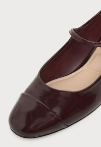 Chaussure Mary Jane en cuir verni bordeaux foncé avec un bout arrondi et une fine bride sur le cou-de-pied, reposant sur un fond clair.