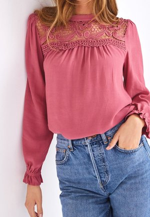 Kvinde iført en lyserød bluse med langt ærme, blondenet på forstykket og pufærmer, kombineret med blå højhalsede jeans, hånd i lommen.