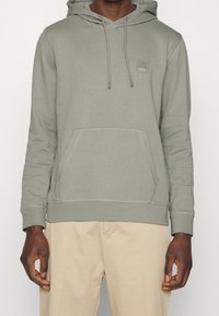 Sudadera con capucha de color verde claro, con un bolsillo frontal tipo canguro, puños acanalados y un pequeño parche de logo en el pecho. Textura de tela suave.