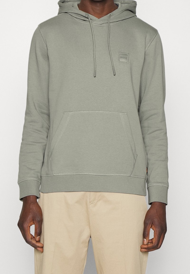 Sudadera con capucha de color verde claro, con un bolsillo frontal tipo canguro, puños acanalados y un pequeño parche de logo en el pecho. Textura de tela suave.