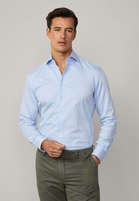 Camicia azzurro chiaro con maniche lunghe e colletto a punta, caratterizzata da una texture liscia e chiusura con bottoni, abbinata a pantaloni verde oliva.