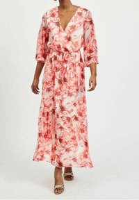 Kvinde iført en lang, flydende hvid kjole med røde blomsterprint, trekvartærmer og V-hals, parret med beige ouvert-toe hæle.