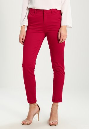 Femme portant un pantalon rouge ajusté, un chemisier blanc à manches longues et des sandales à talons hauts beige, debout devant un fond blanc uni.