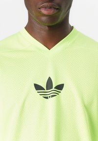 Neon rumena mrežasta športna majica z V-izrezom in črnim Adidas trefoil logotipom na prsih. Lahka in zračna tkanina.