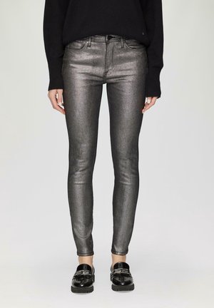 Žena nosí metalicky stříbrné úzké džíny, černý svetr s dlouhými rukávy a černé lesklé mokasíny s řetízkovým detailem, stojí před jednoduchým pozadím.