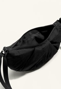 Czarna tkaninowa torba z zamkiem błyskawicznym, posiadająca teksturowaną powierzchnię i regulowany pasek. Zaprojektowana do kompaktowego przechowywania i przenoszenia.