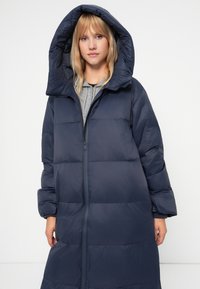 Pufová bunda v tmavom námorníckom odtieni, s prešívaným dizajnom, oversized kapucňou, predným zipsom a rebrovanými manžetami. Materiál sa zdá byť hladký a ľahký.