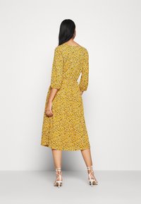Robe midi florale jaune avec des manches trois-quarts, taille ajustée avec ceinture, et jupe fluide. Associée à des talons blancs à lanières.