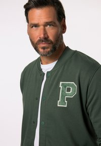 Giacca varsity verde con bottoni, caratterizzata da un grande logo ricamato bianco e verde "P" sul petto sinistro. Camicia bianca sotto.