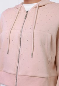 Roza zip-up dukserica s srebrnim ukrasima, koja ima kapuljaču s uže, dva prednja džepa i rebraste manžete i donji rub. Glatka tekstura.