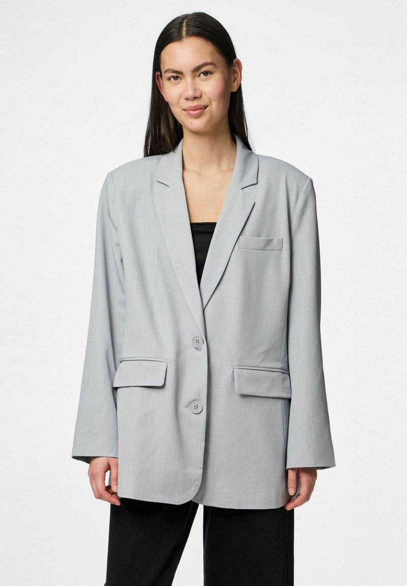 Hele hallikas bleiser, millel on kaks eesmist taskut, ühe nupuga kinnitamine ja lõigatud kraed. Sile tekstuur ja avar lõige, kantud musta särgi peal.