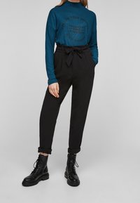 Teal långärmad topp med tryckt text och svarta högmidjade byxor med bälte, kombinerat med svarta snörda kängor.