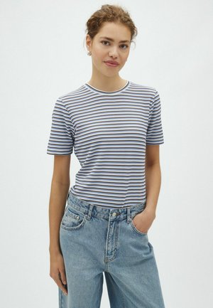 Jeune femme aux cheveux bouclés portant un t-shirt à manches courtes rayé et un jean bleu clair, debout avec une main dans la poche devant un fond blanc.