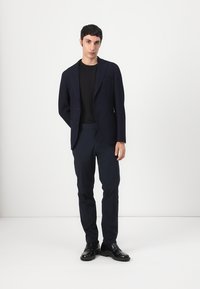 Blazer navy con solapas de muesca, cierre de un solo botón y dos bolsillos frontales, combinado con pantalones oscuros y zapatos de cuero negros.