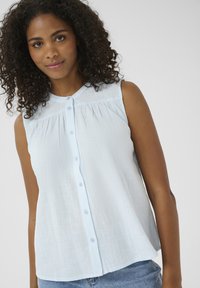 Blouse sans manches bleu clair en tissu texturé, avec un devant boutonné et un détail froncé au niveau du col, associée à un short en denim.
