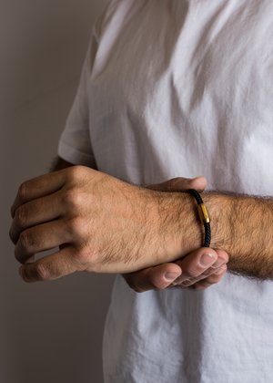 Black woven bracelet with a gold-colored clasp, worn on a wrist, displayed against a white shirt and neutral background.