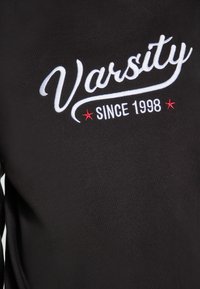 Fekete pulóver, amelyen fehér hímzett felirat található: "Varsity" és "Since 1998" piros kiegészítőkkel, sima textúrával és hosszú ujjakkal.