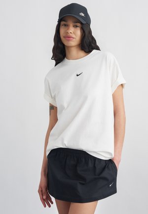 Młoda kobieta ubrana w białą koszulkę Nike, czarną spódnicę i czarną czapkę Nike, stoi z jedną ręką w kieszeni na jasnym tle.