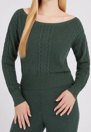 Kobieta ubrana w ciemnozielony dziergany sweter i pasujące spodnie z wzorem warkoczy, pozująca z rękami blisko bioder na jasnym tle.