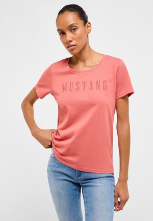 Frau trägt ein korallenrotes Mustang-T-Shirt und hellblaue Jeans, steht mit einer Hand in der Hüfte vor einem schlichten weißen Hintergrund.