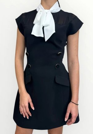 Donna che indossa un vestito nero senza maniche con grandi bottoni e tasche con pattina, accessoriato con una sciarpa bianca al collo.