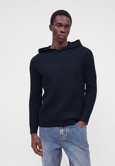 Jeune homme aux cheveux courts portant un sweat à capuche noir texturé et un jean bleu clair, debout avec une main dans la poche devant un fond uni.