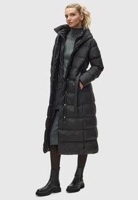 Lange zwarte pufferjas met een capuchon, drukknopen, twee pockets en een strikceintuur, gedragen over een grijze jurk en zwarte enkelboots.