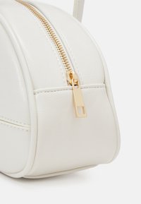 Coin d'un sac à main en cuir texturé blanc avec tirette de fermeture éclair en métal doré et détails de couture sur un fond clair.