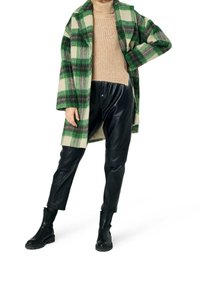 Cappotto di lana a quadri verde e beige sopra un maglione di filo beige, abbinato a pantaloni di pelle nera e stivaletti neri.