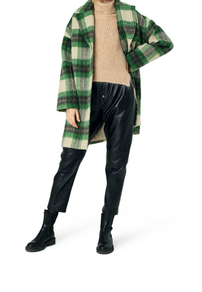 Cappotto di lana a quadri verde e beige sopra un maglione di filo beige, abbinato a pantaloni di pelle nera e stivaletti neri.