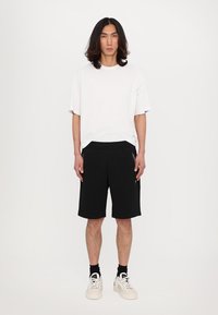 Hvit bomull T-skjorte kombinert med svarte shorts. Shortsen har to glidelås-lommer og en elastisk midje. Modellen har på seg hvite joggesko.