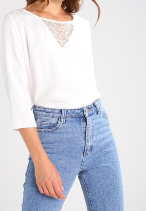 Witte longsleeve top met een kanten driehoekdetail bij de halslijn, gecombineerd met lichtblauwe high-waisted spijkerbroek.