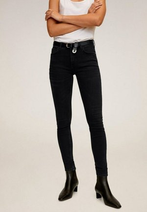 Personne portant un jean skinny noir, des bottines noires à talons et un top blanc sans manches, debout les bras croisés.