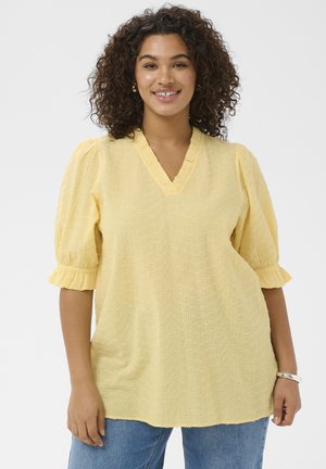 Uśmiechnięta kobieta o kręconych włosach, ubrana w żółtą, fakturowaną bluzkę z marszczonymi rękawami i niebieskie dżinsy, stojąca na białym tle.