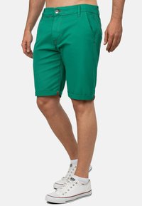 Pantaloni corti in cotone verde con una vestibilità slim, orli arrotolati e cuciture rosse a contrasto. Indossati con sneakers bianche con accenti rossi.