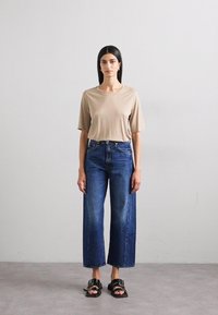 Beige T-shirt met korte mouwen, hoge taille wijd uitlopende donkere spijkerbroek en zwarte sandalen met gespen, weergegeven tegen een lichtgrijze achtergrond.