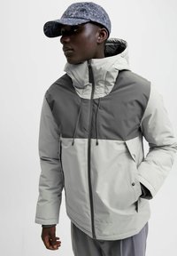 Hellegraue Pufferjacke mit dunkelgrauen Akzenten, hohem Kragen, verstellbarem Kordelzugkapuze und Seitentaschen. Passt gut zu einer gemusterten Mütze.