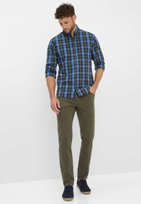 Karohemd mit blauem, grünem und schwarzem Muster, Knopfdesign, lange Ärmel bis zu den Ellenbogen gerollt. Olivgrüne Hose und marineblaue Sneakers.
