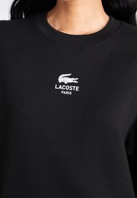 Felpa nera in cotone, con logo bianco Lacoste e la scritta "PARIS" sul petto, colletto rotondo e una texture liscia.