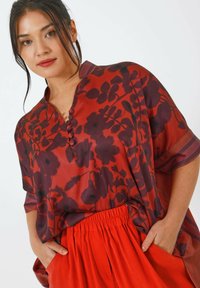 Blouse fluide à fleurs rouge-violet avec un col en V, cinq boutons et des manches courtes. Associée à une jupe rouge vif à taille élastique.