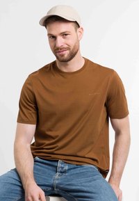 Camiseta marrón de manga corta con cuello redondo, que presenta un pequeño logo. El modelo lleva una gorra color beige claro y jeans de mezclilla.