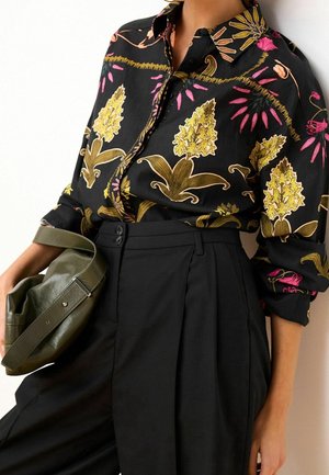 Kobieta w czarnej koszuli na guziki z żółto-różowym kwiatowym wzorem, czarnych plisowanych spodniach z wysokim stanem, trzymająca oliwkowo-zieloną skórzaną torbę.