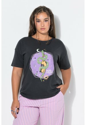 Kobieta z długimi włosami nosi czarny T-shirt z zielonym wężem i fioletowym wzorem oraz różowe spodnie w paski, stoi na białym tle.