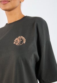 Donkergrijze katoenen t-shirt met een rond grafisch ontwerp in licht perzik, met de tekst "WEES VRIENDELIJK VOOR JE GEEST" en een zonmotief.