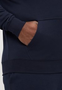 Σκούρος μπλε φούτερ με ελαστικές μανσέτες και στρώση κάτω, διαθέτει μπροστινή τσέπη και μαλακό, υφασμάτινο υλικό. Κοντινή λήψη χεριού στην τσέπη.