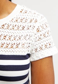 Mangas cortas de encaje de crochet blanco con patrón floral, combinadas con un cuerpo de jersey a rayas blancas y azul marino. Textura suave y diseño ajustado.