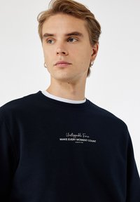 Joven con cabello rubio y aros en las orejas, vistiendo una sudadera negra con el texto "Fuerza Imparable HAZ QUE CADA MOMENTO CUENTE", mirando de lado.