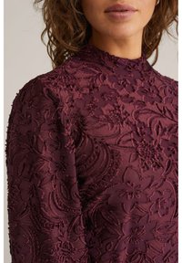 Bordinės spalvos suknelė su tekstūruotu gėlių siuvinėjimu, aptempto silueto, aukšta apykakle ir ilgomis rankovėmis, atskleidžiančiomis sudėtingas detales.