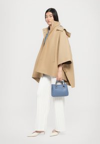 Manteau beige en style poncho avec des boutons pression, superposé à un top bleu, associé à un pantalon blanc plissé et un petit sac à main bleu.