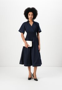 Vestido de corte envolvente azul-marinho com mangas curtas, cintura franzida e costura em contraste. Bolsa branca a tiracolo e sapatos pretos de salto alto realçam o visual.